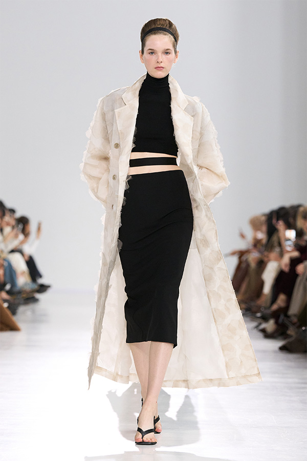 Défilé Max Mara Printemps Été 2026 : look avec trench-coat clair sur col roulé et jupe coupe ajustée noirs.